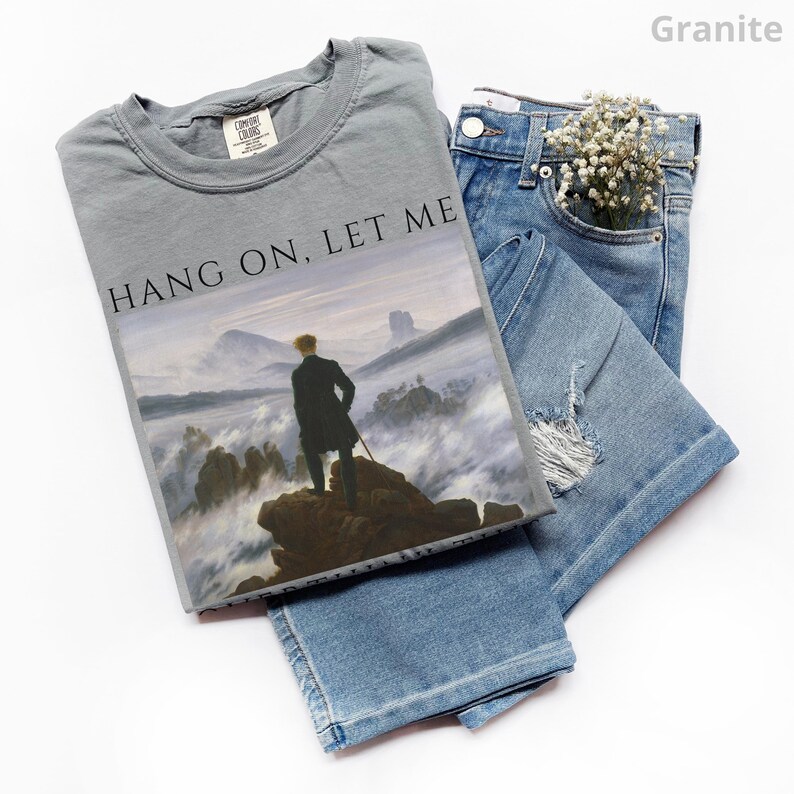 May include: A gray t-shirt with the text "HANG ON, LET ME" printed on the front. The shirt features a painting of a man standing on a mountaintop with fog in the background.
