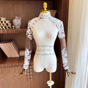 Puede incluir: Un maniquí de costura color crema con cuello alto y mangas largas. Las mangas son transparentes, con detalles de encaje floral blanco. El maniquí está sobre un soporte dorado. El texto "DanielPHCo" está impreso.