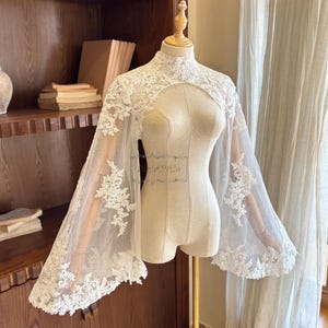 May include: A white lace bridal cape with a high neckline and long, sheer sleeves. The cape features intricate floral lace detailing along the neckline and sleeve edges. The garment is displayed on a mannequin.