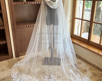 May include: A sheer white bridal cape with a hood, displayed on a mannequin. The cape features a delicate floral lace trim along the hood and hem. The cape is long, flowing, and ideal for a wedding or special occasion.
