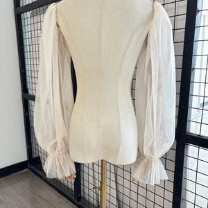 May include: A white sheer blouse with long, puffed sleeves and ruffled cuffs. The blouse is on a white dress form.