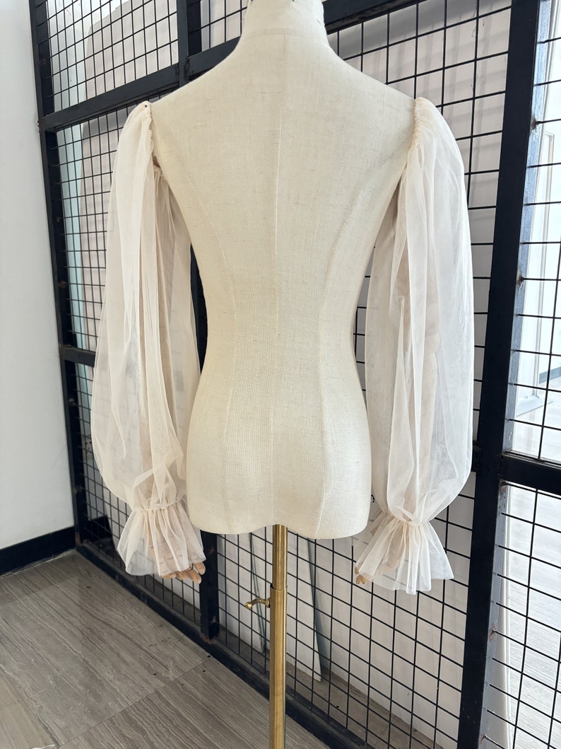May include: A white sheer blouse with long, puffed sleeves and ruffled cuffs. The blouse is on a white dress form.