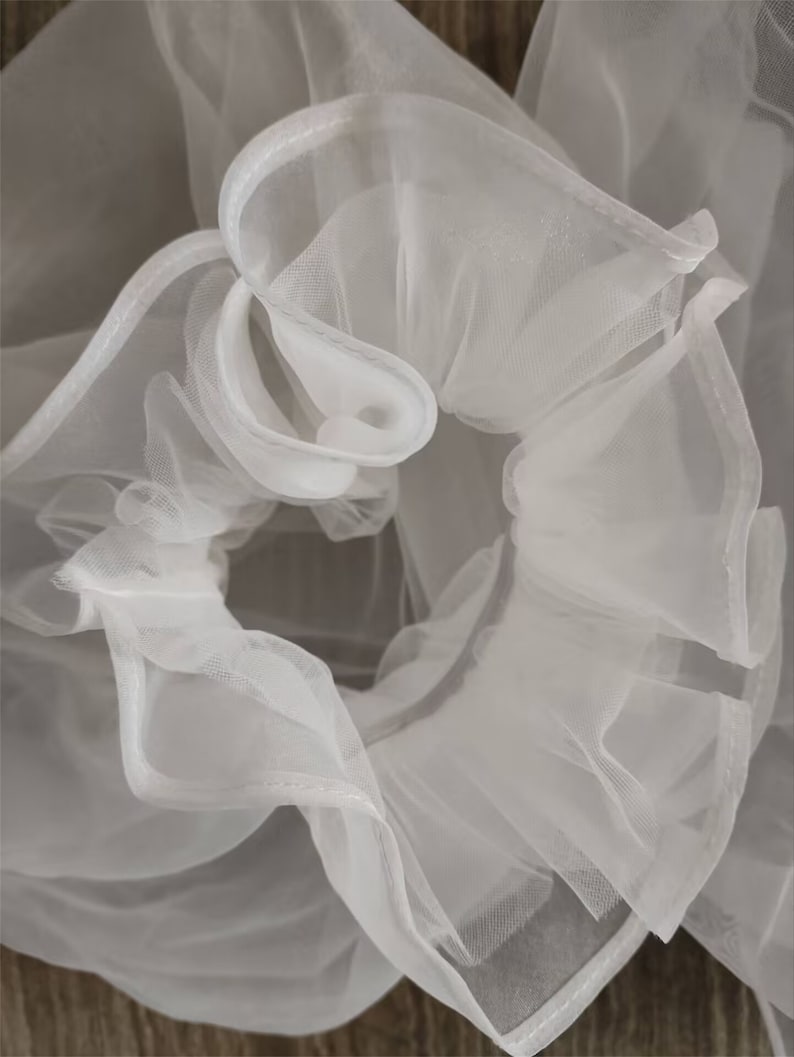 May include: White tulle fabric with a ruffled edge. The fabric is soft and delicate, and it has a slightly sheer appearance.