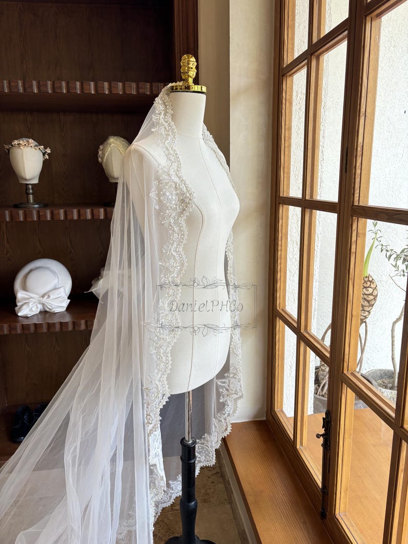 Puede incluir: Un velo de novia blanco con un borde de encaje con cuentas. El velo est&aacute; drapeado sobre un maniqu&iacute; blanco y se exhibe frente a una ventana.