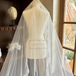 Puede incluir: Un velo de novia blanco con un delicado borde de encaje y un borde de cuentas. El velo est&aacute; drapeado sobre un maniqu&iacute; y se muestra sobre un fondo de madera.