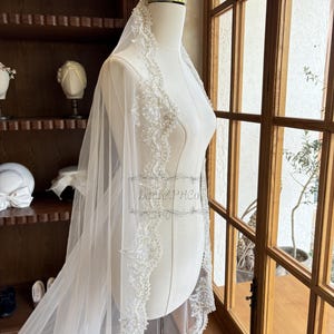 Puede incluir: Un velo de novia blanco con un borde de encaje con cuentas. El velo est&aacute; drapeado sobre un maniqu&iacute; blanco y se exhibe frente a una ventana.
