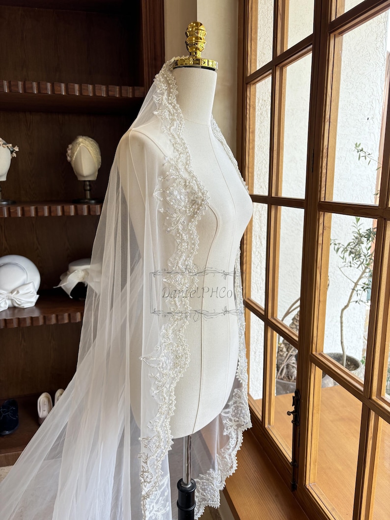 Puede incluir: Un velo de novia blanco con un borde de encaje con cuentas. El velo est&aacute; drapeado sobre un maniqu&iacute; blanco y se exhibe frente a una ventana.