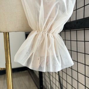 May include: Close-up of a white sheer fabric with a ruffled edge. The fabric is gathered at the wrist and has a delicate, airy feel.