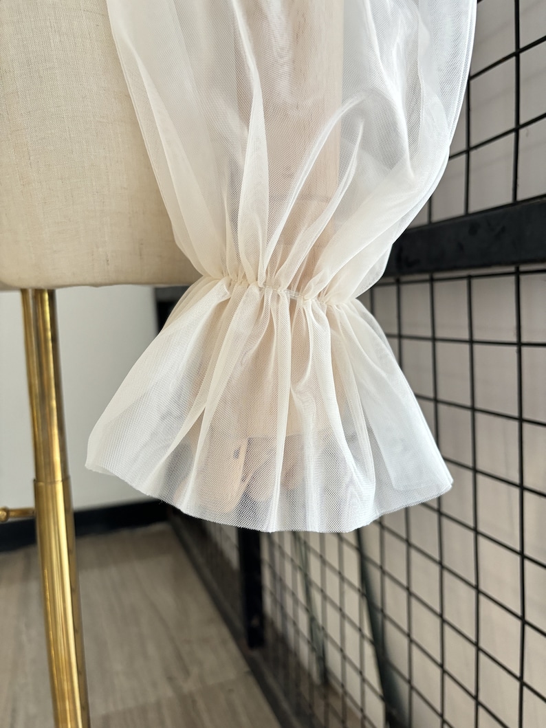 May include: Close-up of a white sheer fabric with a ruffled edge. The fabric is gathered at the wrist and has a delicate, airy feel.