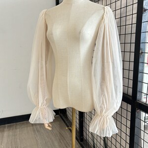 May include: A sheer white blouse with long, puffed sleeves and ruffled cuffs. The blouse is on a white mannequin.