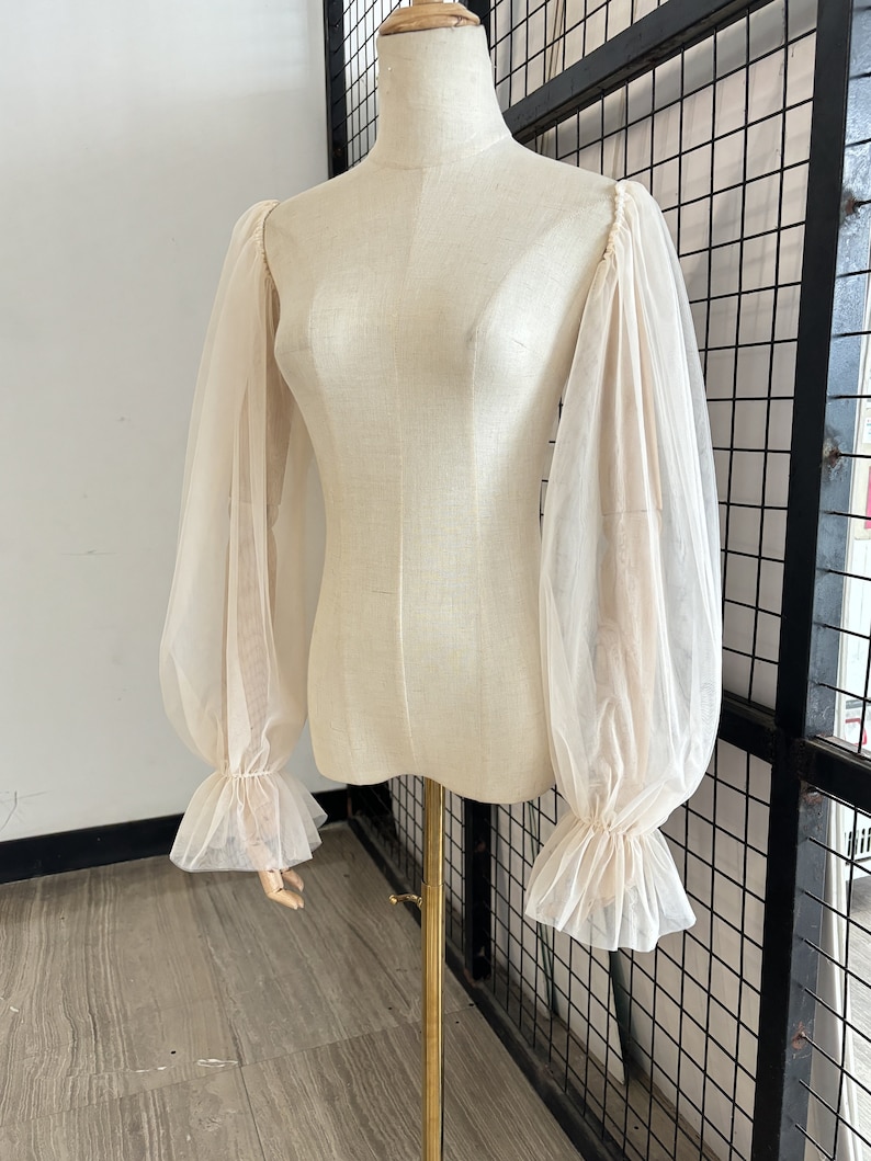 May include: A sheer white blouse with long, puffed sleeves and ruffled cuffs. The blouse is on a white mannequin.