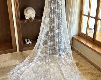 May include: A long, white lace veil with a floral pattern, displayed on a mannequin. The veil cascades down, showcasing its intricate design. The image also features a wooden bookshelf with hats and a window with natural light.