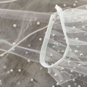 Peut inclure: Tissu de tulle blanc avec des perles &eacute;parpill&eacute;es. Le tissu est rassembl&eacute; et pli&eacute;, cr&eacute;ant une texture douce et fluide.