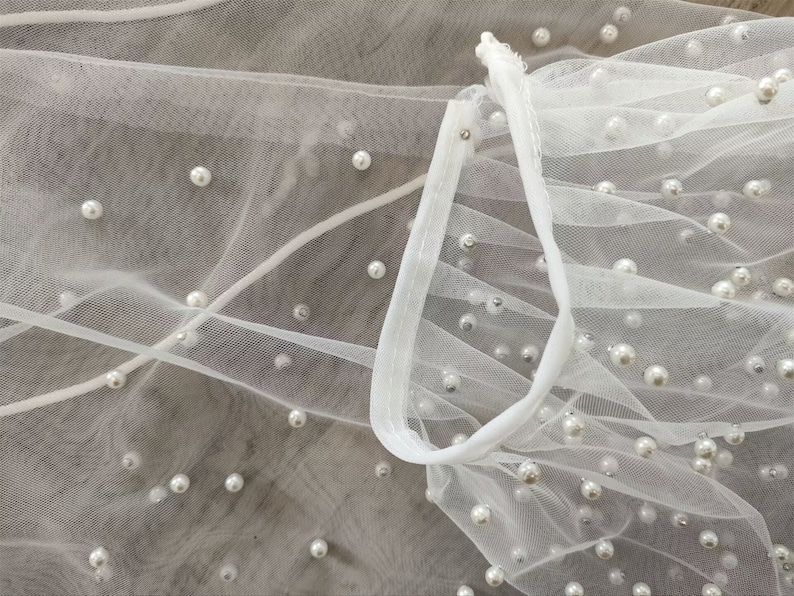 Peut inclure: Tissu de tulle blanc avec des perles &eacute;parpill&eacute;es. Le tissu est rassembl&eacute; et pli&eacute;, cr&eacute;ant une texture douce et fluide.