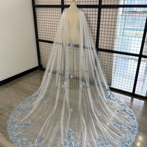 May include: A white tulle bridal cape with a deep V-neckline and a delicate floral design in light blue. The cape is shown on a dress form against a black metal grid background.