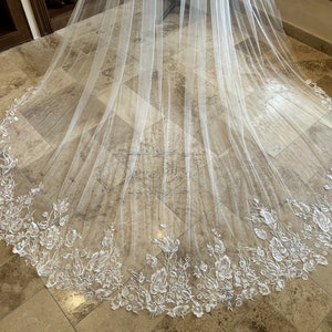 May include: A white tulle wedding veil with a delicate floral lace trim. The veil is laid out on a tiled floor.