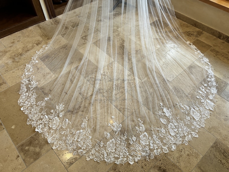 May include: A white tulle wedding veil with a delicate floral lace trim. The veil is laid out on a tiled floor.