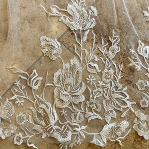 May include: Close-up of a white lace fabric with a floral pattern. The lace is delicate and intricate, with small flowers and leaves.