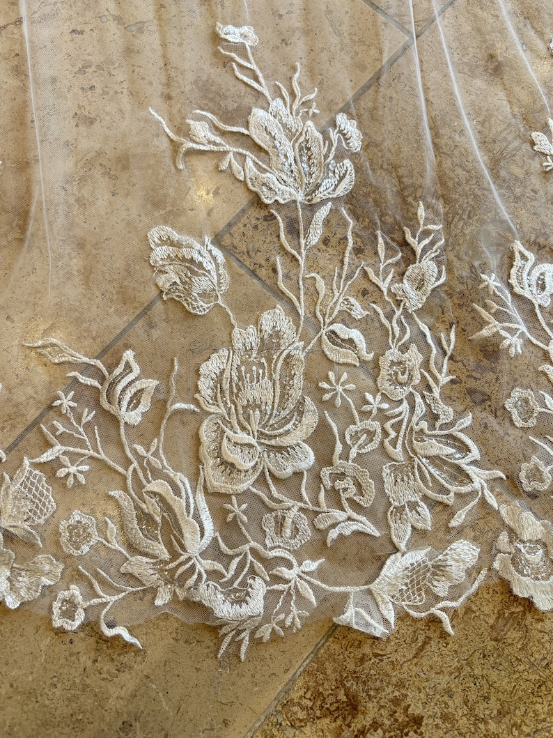 May include: Close-up of a white lace fabric with a floral pattern. The lace is delicate and intricate, with small flowers and leaves.