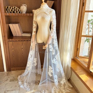 Könnte beinhalten: Ein weißes Braut-Cape und Bolero-Set aus Spitze, präsentiert auf einer Schaufensterpuppe. Der Bolero hat lange Ärmel mit ausgestellten Manschetten und einen hohen Ausschnitt. Das Cape reicht bis zum Boden und hat aufwendige florale Spitzenverzierungen.