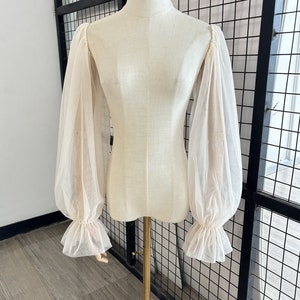 May include: A sheer, off-white blouse with long, gathered sleeves and ruffled cuffs. The blouse is on a white mannequin.