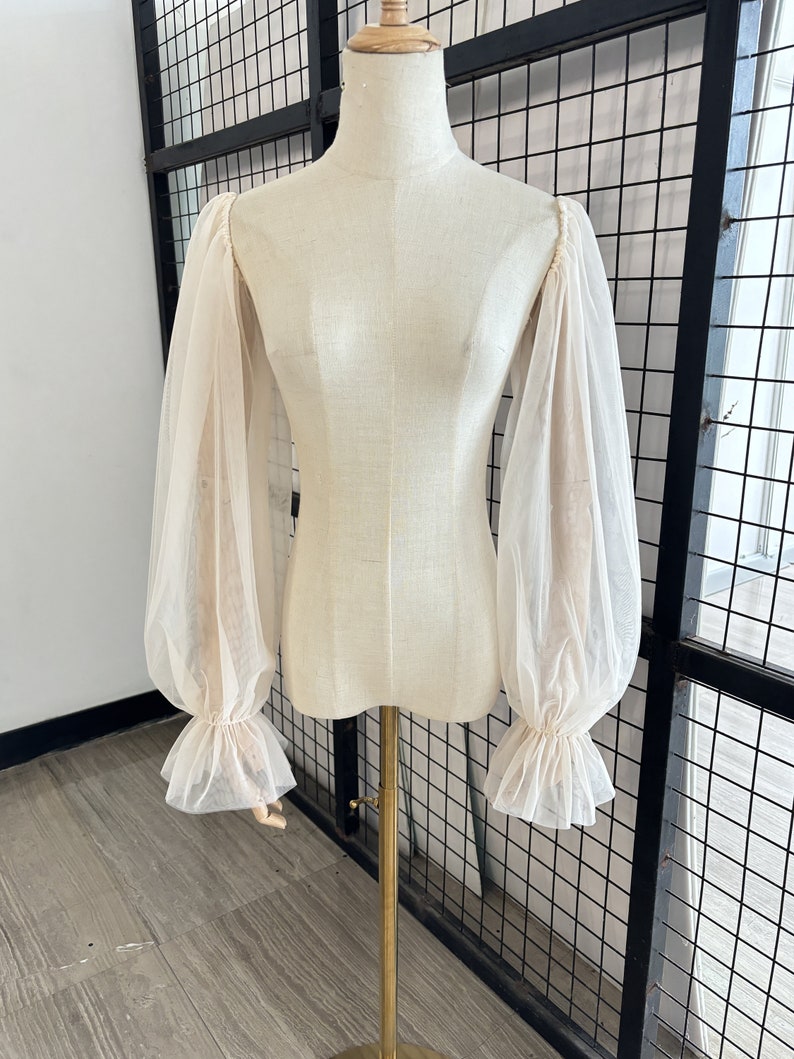 May include: A sheer, off-white blouse with long, gathered sleeves and ruffled cuffs. The blouse is on a white mannequin.
