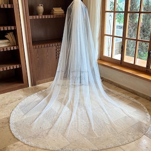 May include: A long, white bridal veil with a beaded edge and scattered pearl accents. The veil is displayed on a mannequin, showcasing its flowing design. The veil is made of a sheer fabric.