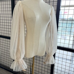 May include: A white sheer blouse with long, puffed sleeves and ruffled cuffs. The blouse is on a white mannequin.