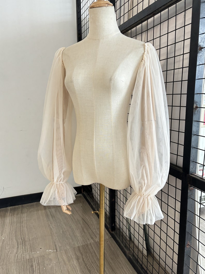 May include: A sheer white blouse with long, puffed sleeves and ruffled cuffs. The blouse is on a white mannequin.