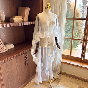 Op de afbeelding: Een doorschijnende witte bruidssluier met delicate kanten rand is over een mannequin gedrapeerd. De sluier wordt op een goudkleurige standaard voor een raam getoond. De mannequin heeft houten armen en een goudkleurige kop.