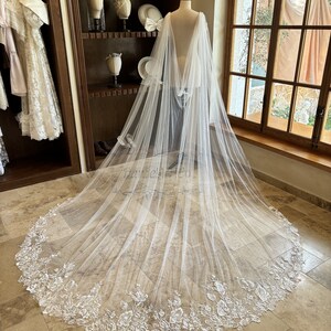 May include: A white tulle bridal veil with a floral lace trim. The veil is draped over a mannequin and has a long, flowing train.