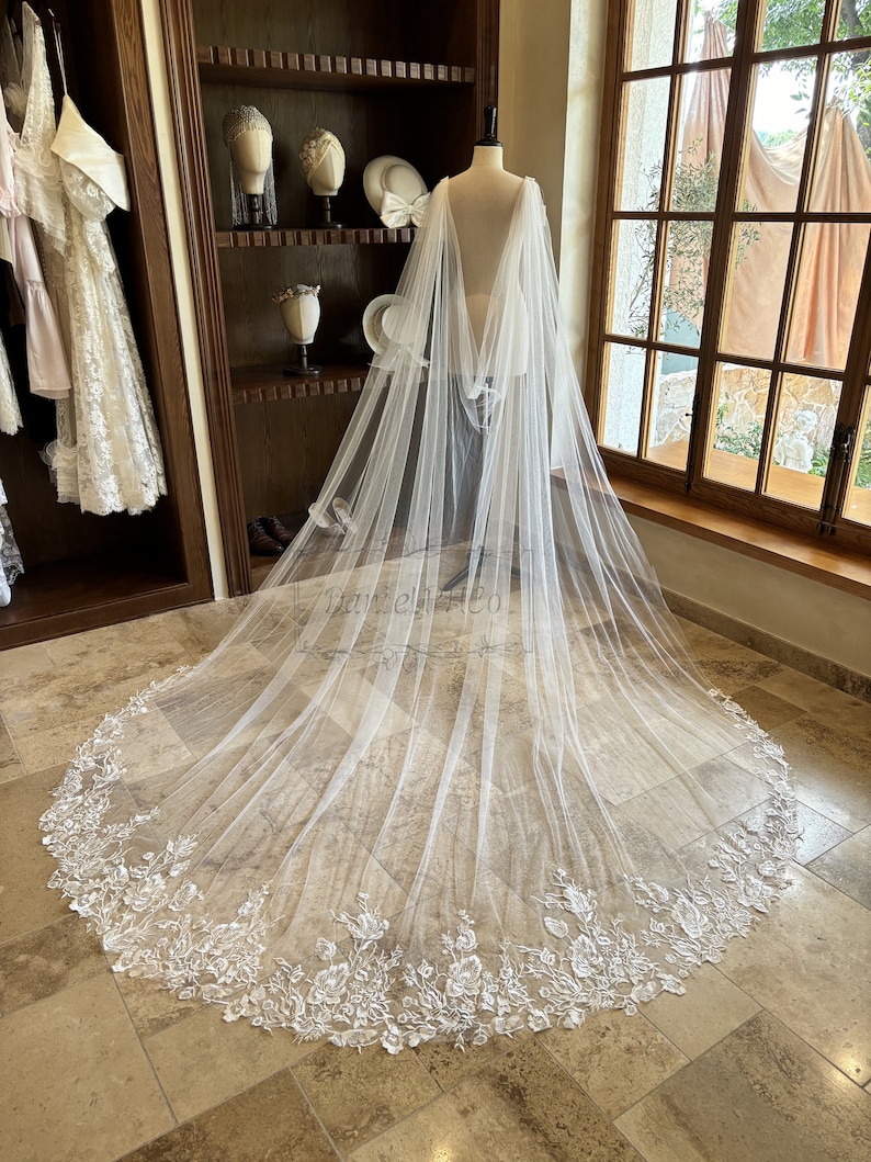 May include: A white tulle bridal veil with a floral lace trim. The veil is draped over a mannequin and has a long, flowing train.