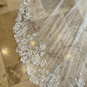 May include: A white bridal veil featuring delicate floral lace embroidery. The veil is made from soft tulle and has a scalloped edge.