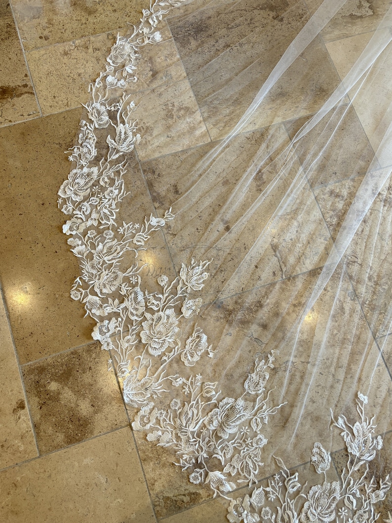 May include: A white bridal veil featuring delicate floral lace embroidery. The veil is made from soft tulle and has a scalloped edge.