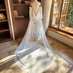 May include: A white wedding dress with a long train is displayed on a mannequin. The dress features off-the-shoulder sleeves and a draped back. The text "Daniel PH Co" is visible on the train. The dress is in front of a window and a wooden bookshelf.