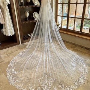 Könnte beinhalten: Ein weißer Braut Schleier mit einer zarten floralen Spitzenborte. Der Schleier ist lang und fließend, mit einer weichen, durchsichtigen Textur.