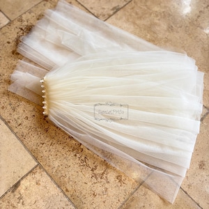 May include: Ivory-colored bridal veil made of layered tulle. The veil is gathered at the top and attached to a comb. The veil is displayed on a light brown tiled surface. The text "Daniel PH Co" is visible on the veil.