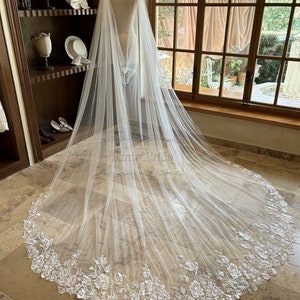 May include: A white tulle bridal veil with a scalloped edge and floral embroidery. The veil is draped over a mannequin and is lying on a tiled floor.