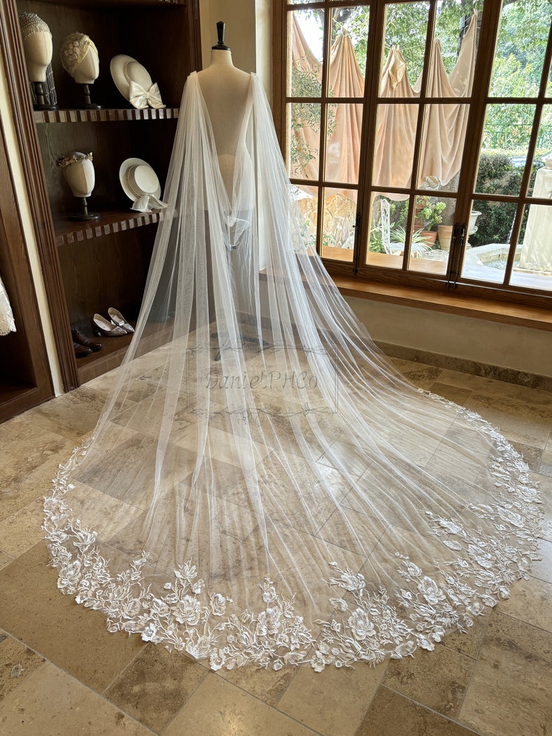 May include: A white tulle bridal veil with a scalloped edge and floral embroidery. The veil is draped over a mannequin and is lying on a tiled floor.