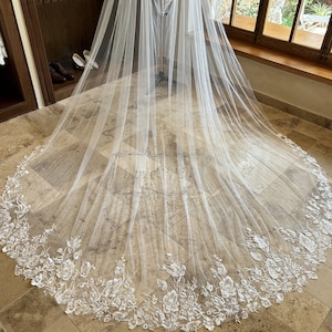 May include: A white tulle wedding veil with a floral lace trim. The veil is draped over a mannequin and is displayed on a tiled floor.