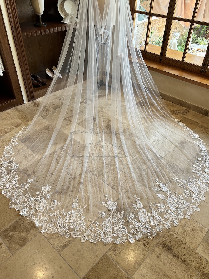 May include: A white tulle wedding veil with a floral lace trim. The veil is draped over a mannequin and is displayed on a tiled floor.