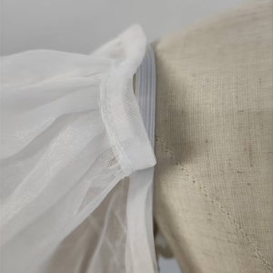 May include: Close-up of a white tulle fabric with a white elastic band sewn along the edge. The fabric is gathered and pleated, creating a soft and delicate look.