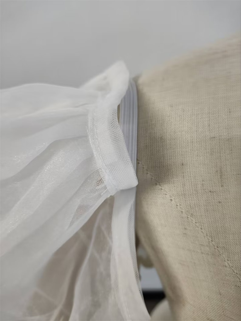 May include: Close-up of a white tulle fabric with a white elastic band sewn along the edge. The fabric is gathered and pleated, creating a soft and delicate look.