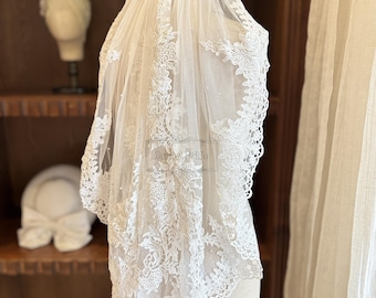 May include: A delicate, ivory-colored bridal veil with intricate floral lace detailing. The veil is draped over a mannequin, showcasing its flowing design and scalloped edges. The lace pattern is visible throughout the veil.