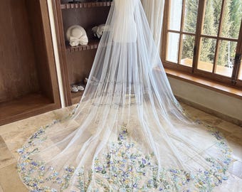 May include: A white bridal veil with a floral design. The veil is made of tulle and features embroidered flowers in shades of blue, yellow, and green. The veil is long and flowing, and it would be perfect for a wedding.