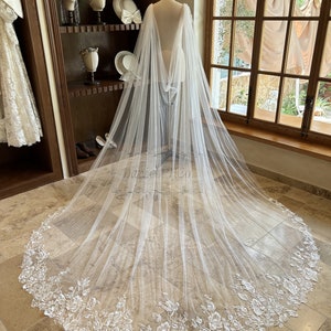 May include: A white tulle bridal veil with a floral lace trim. The veil is draped over a mannequin and has a long train.