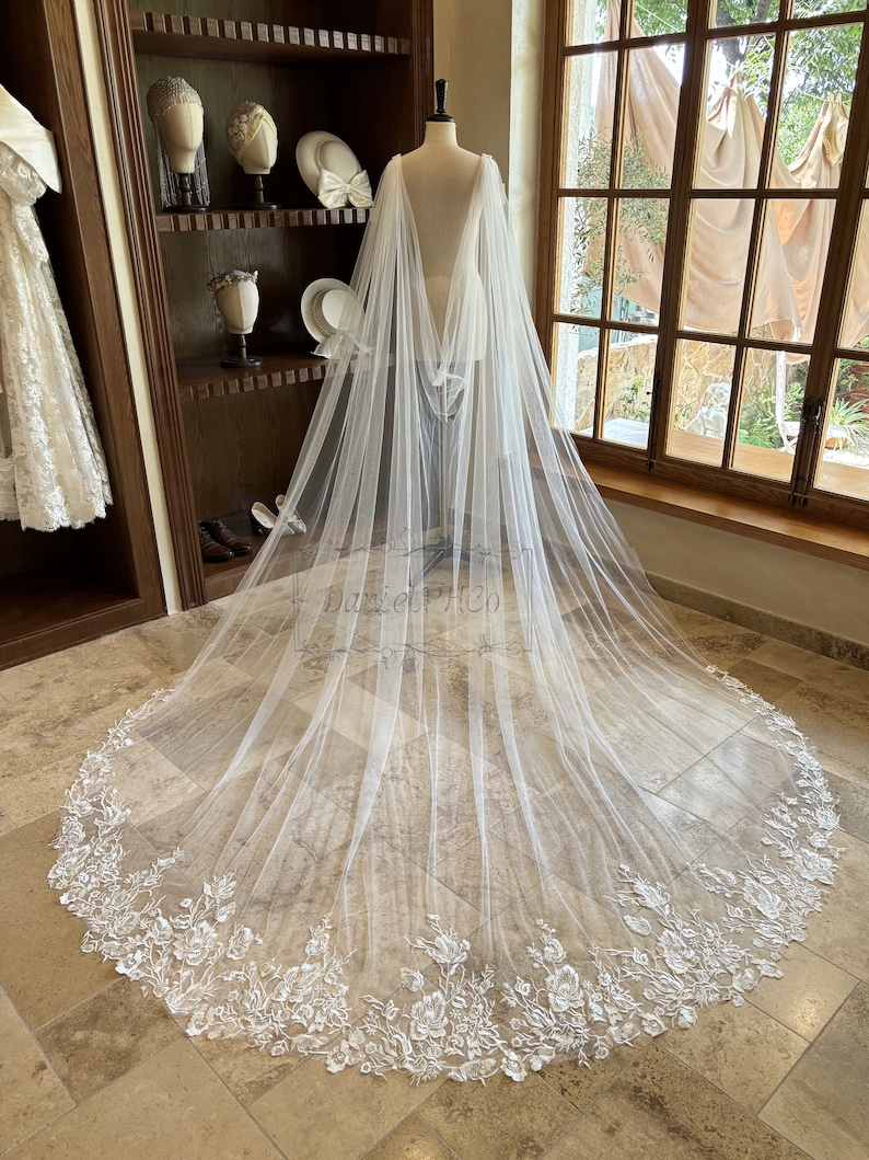 May include: A white tulle bridal veil with a floral lace trim. The veil is draped over a mannequin and has a long train.