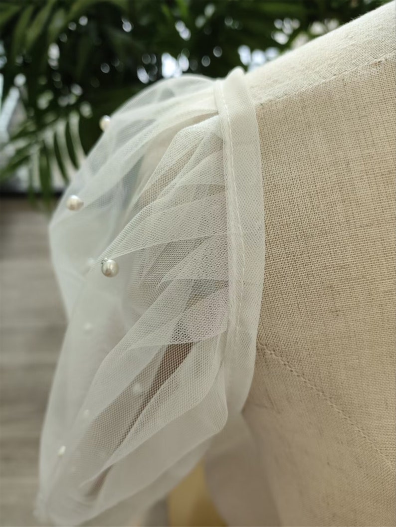 Peut inclure: Tissu de tulle blanc avec des embellissements de perles. Le tulle est fronc&eacute; et pliss&eacute;, cr&eacute;ant un look d&eacute;licat et f&eacute;minin.