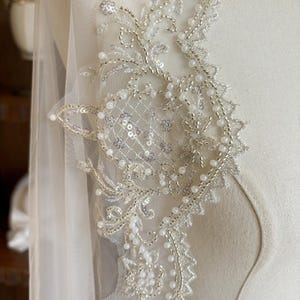 Puede incluir: Primer plano de una delicada tela de encaje de color marfil con un dise&ntilde;o floral. El encaje est&aacute; adornado con peque&ntilde;as cuentas blancas y lentejuelas, creando un efecto brillante.