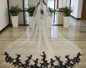 May include: A long white tulle wedding veil with a black floral lace trim. The veil is draped over a mannequin and is shown from the back.